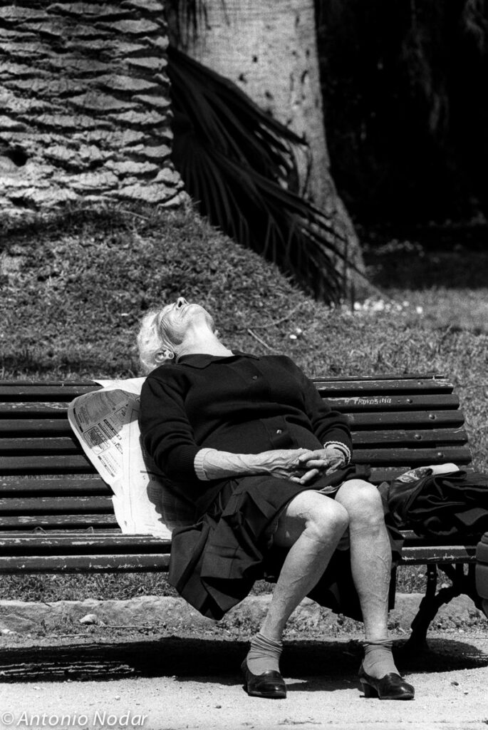 Elderly woman leans back on a bench, face tilted toward the sun, newspaper and handbag beside her; palm tree and park background, Barcelona, 1990s.
