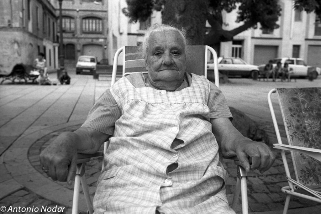 Elderly woman seated on a folding chair in a Barcelona plaza, blurred face, relaxed posture, surrounded by cars and distant groups.