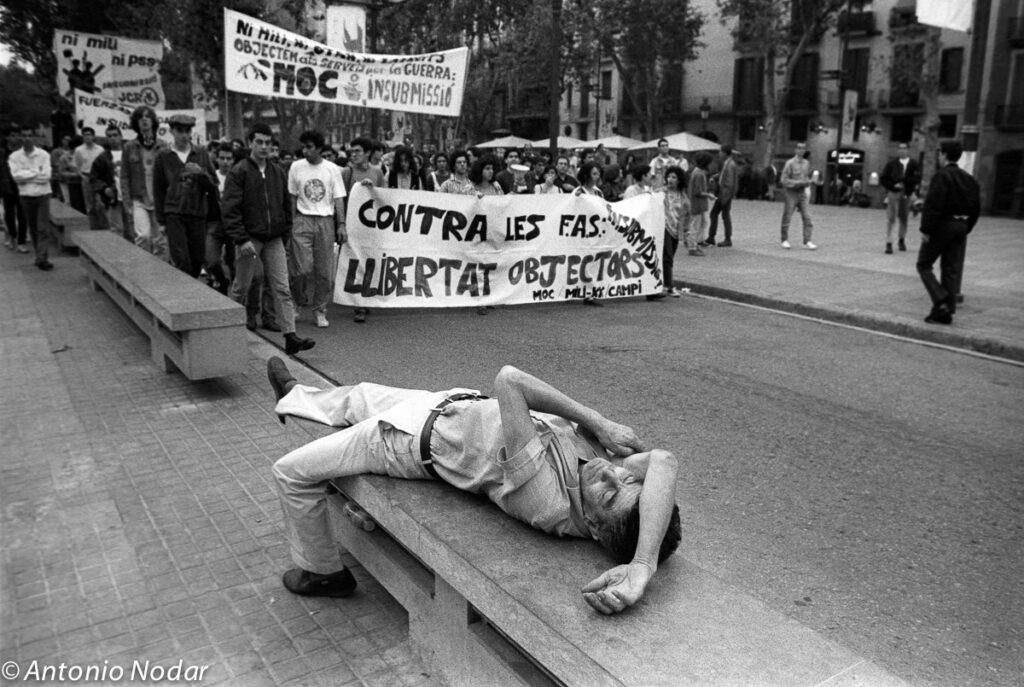 Protesters march in Barcelona holding banners, while a man reclines on a stone bench, eyes half closed, street scene, 1990s.