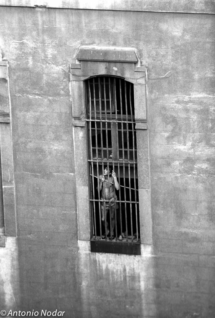 Shirtless man stands behind bars of a tall window in a weathered wall, looking outward, Barcelona, 1990s.
