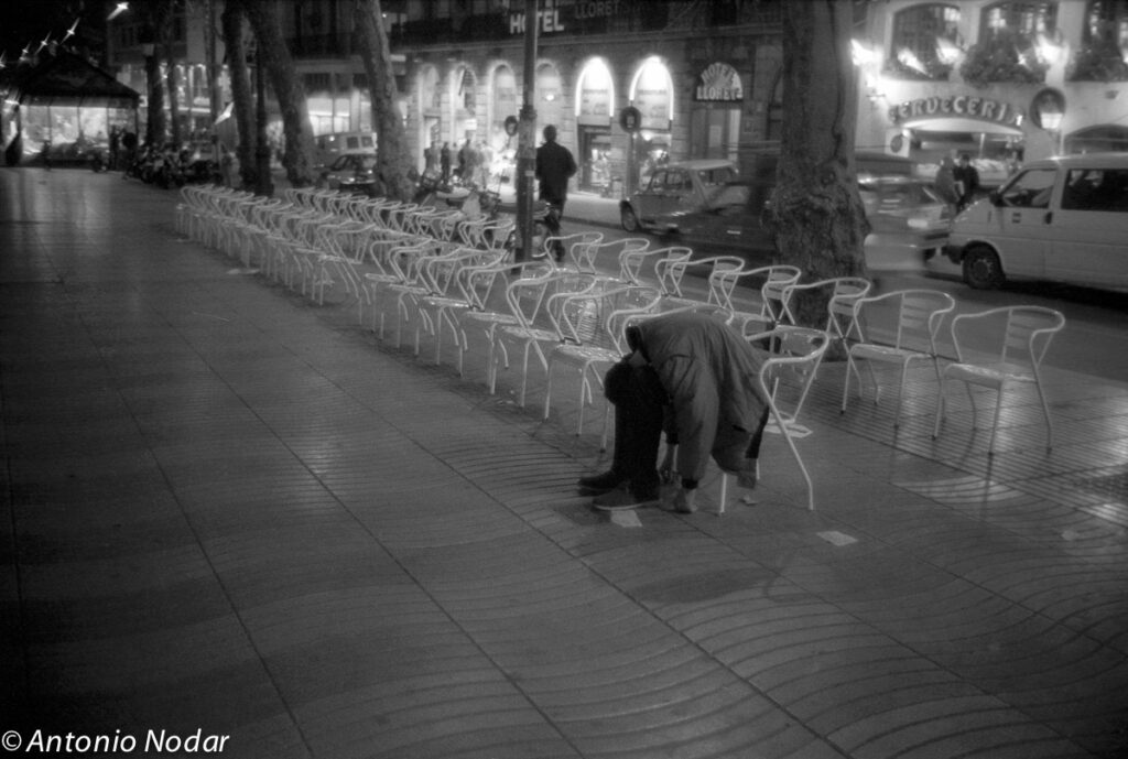Person slumped forward on a metal chair among rows of empty chairs on a lit Barcelona boulevard at night, cars and hotel visible.