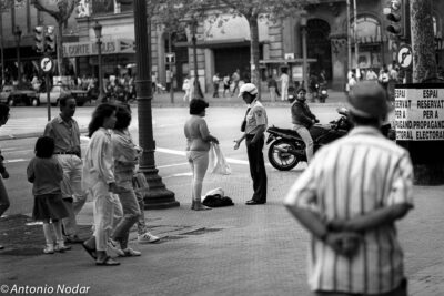 Shirtless woman and police officer interact at a busy street corner in Barcelona while pedestrians, traffic, and a motorbike form the backdrop, 1990s.