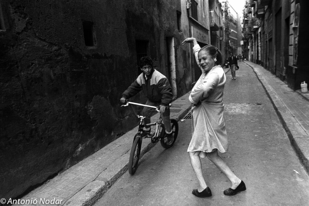 Woman in a dress performs a dramatic gesture as a boy on a bicycle rides by in a narrow Barcelona street, 1990s.