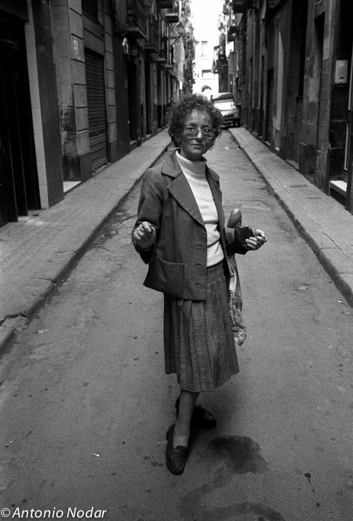 Middle-aged woman stands in the middle of a narrow street holding bread and shopping, turned toward the camera, Barcelona, 1990s.