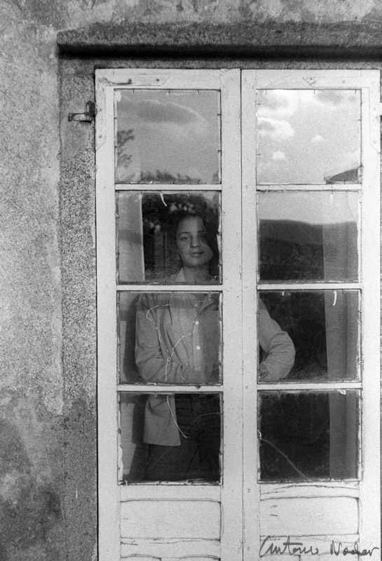 A person stands behind a glass-paned door, partially obscured by reflections, inside a rustic building with stone walls visible outside.