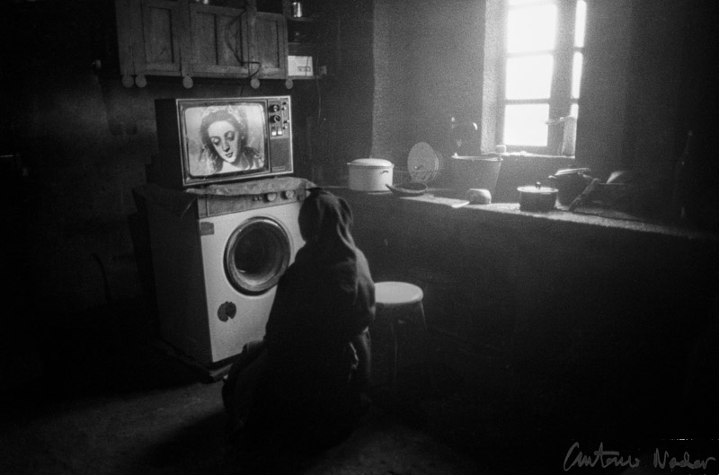 A person in traditional attire sits on a low stool in a dimly lit farmhouse kitchen, intently watching a television set placed on top of a washing machine with sunlight streaming through the window.