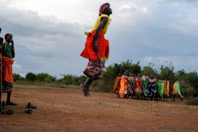 Samburu warrior jumps high into the air barefoot, clad in vibrant traditional attire, with a group of women observing the Adumu dance in Kenya’s Rift Valley