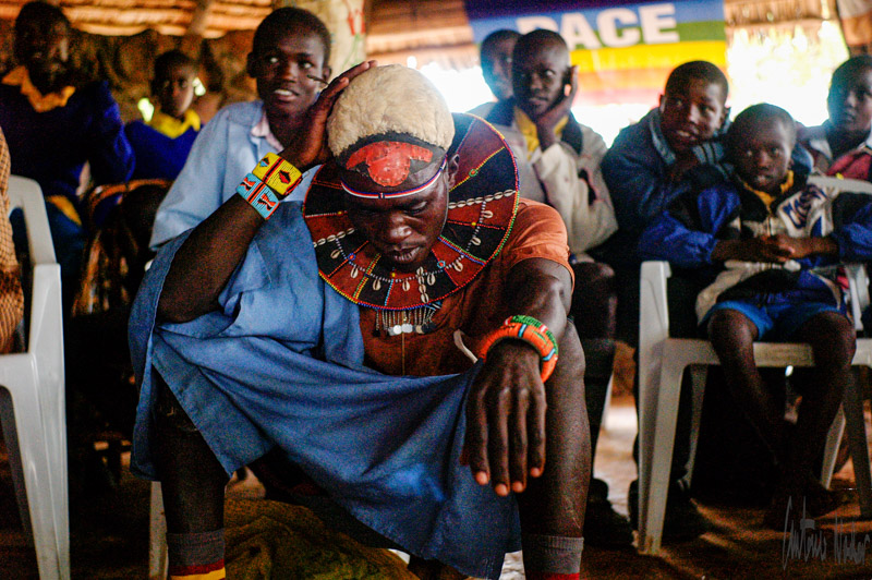 A person dressed in traditional Samburu beadwork and vibrant clothing is seated with one arm resting on their knee among a group gathered for a community event, surrounded by others in similar attire on white chairs.