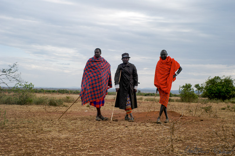 Three Samburu elders dressed in traditional shukas and long coats, standing with walking sticks on the arid plains of northern Kenya beneath a cloudy sky