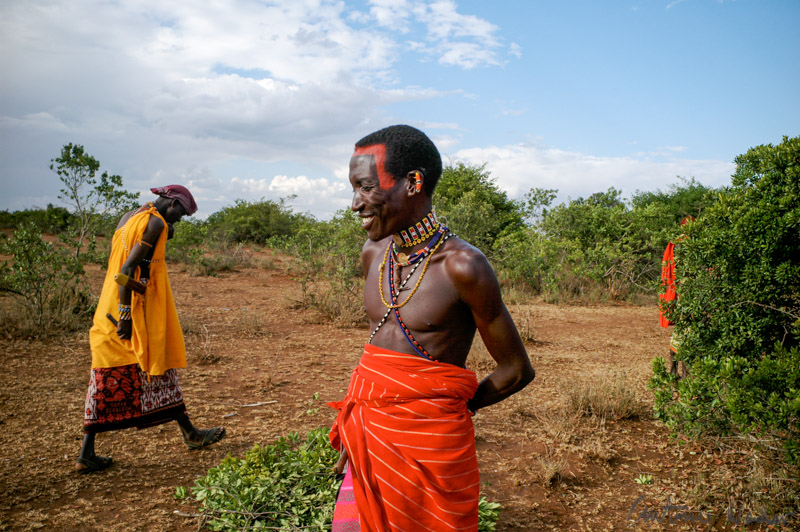 Samburu warrior clad in a red shuka and adorned with bead necklaces stands in the Kenyan bush, with another figure in yellow clothing walking nearby