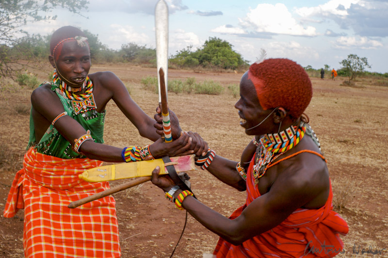 Two Samburu warriors, dressed in colorful beadwork and patterned shukas, engage in a traditional stick fighting demonstration on the dry plains of Kenya’s Rift Valley