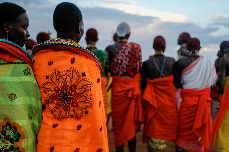 Samburu women adorned in bright orange and green floral shawls and bead collars stand together during a traditional gathering at sunset in Kenya’s Rift Valley