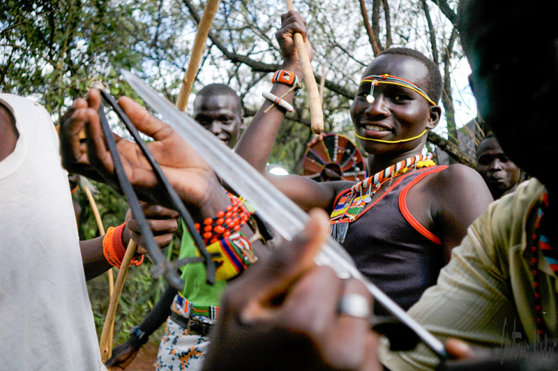 Young men in colorful beadwork and traditional dress gather outside, handling bows and arrows amid the trees, demonstrating techniques and tools used by their ancestors.