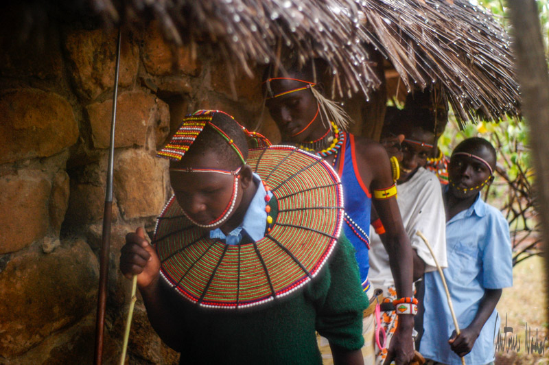 A young person wears vibrant Samburu beadwork outside a thatched hut, accompanied by peers during a cultural heritage gathering in Laikipia.