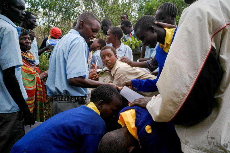 A group of students in blue and yellow uniforms crowd around a tree and an elder holding a twig, writing observations in their notebooks.