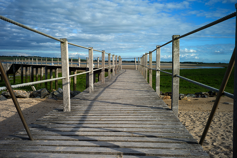 A long wooden boardwalk with rope railings stretches across sand and marshland beneath a partly cloudy sky in Normandy.