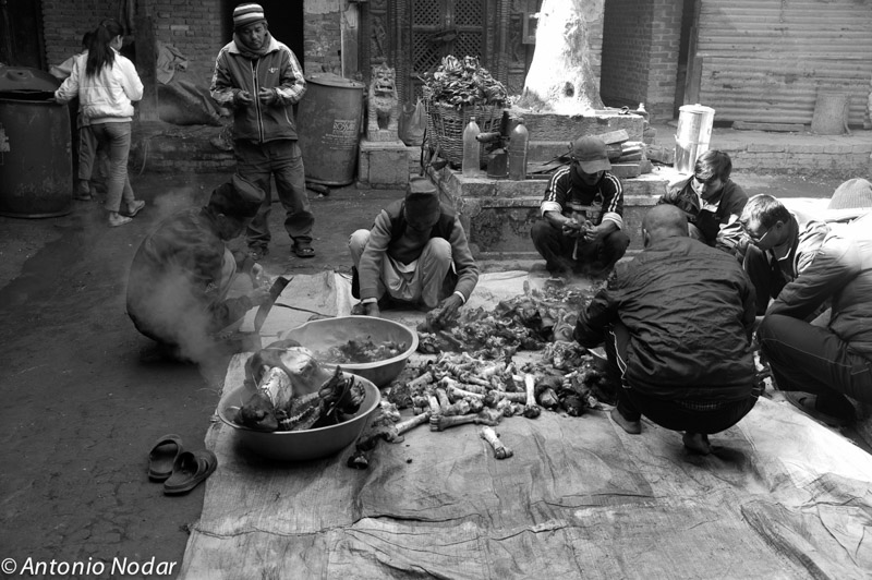 A group of men gathers around steaming bowls and piles of meat parts, preparing food in a Bungamati street, Nepal