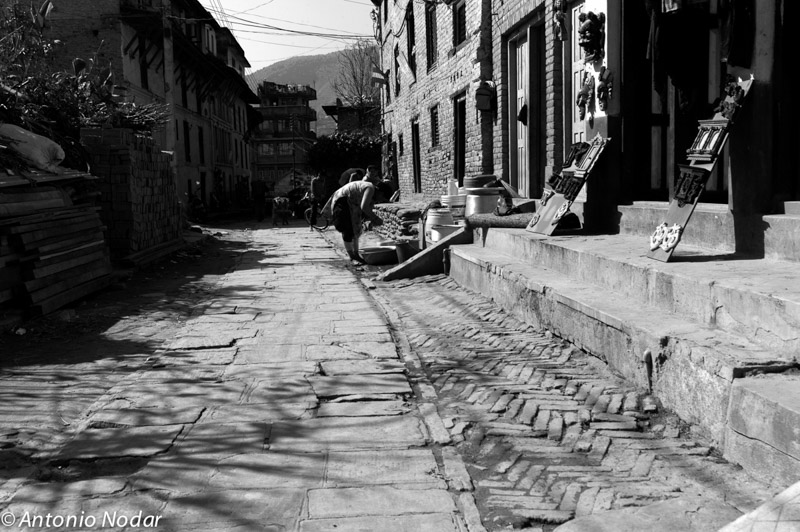 A man bends over while washing or tending items beside brick buildings and crafts displayed outside shops in Bungamati, Nepal