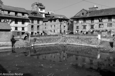 A reflective pond is surrounded by sloping staircases and old brick houses with laundry hanging from windows in Bungamati, Nepal