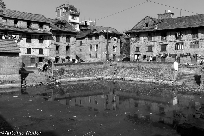 A reflective pond is surrounded by sloping staircases and old brick houses with laundry hanging from windows in Bungamati, Nepal