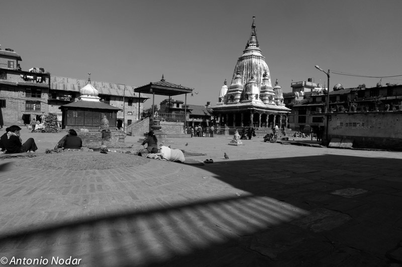 Wide temple square in Bungamati, Nepal, with locals sitting and walking, dominated by a large white temple and historic architecture under clear sky
