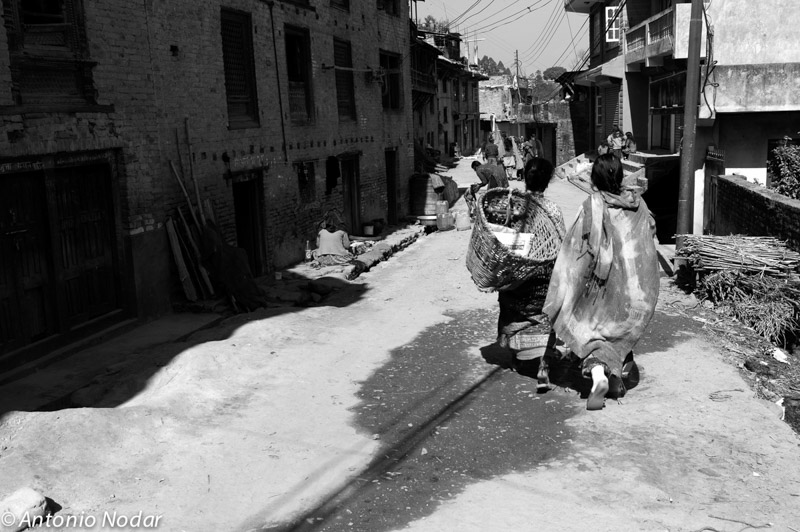 Two women, seen from behind, walk down a sunlit street in Bungamati, Nepal, carrying baskets and wearing traditional attire