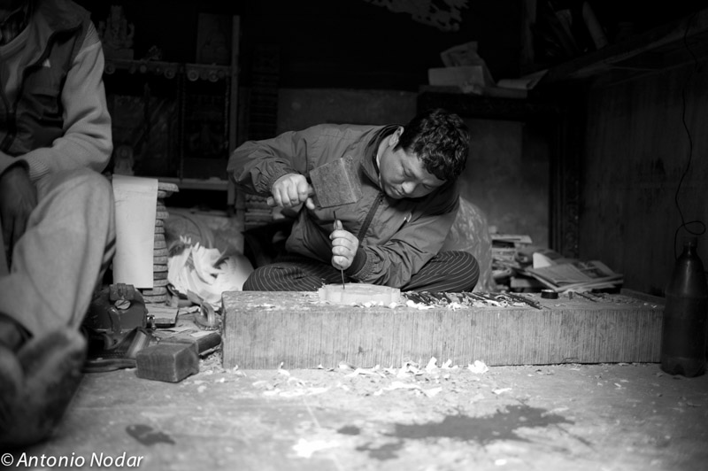A craftsman uses a mallet and chisel to carve wood in a dimly lit Bungamati workshop, surrounded by wood shavings and tools