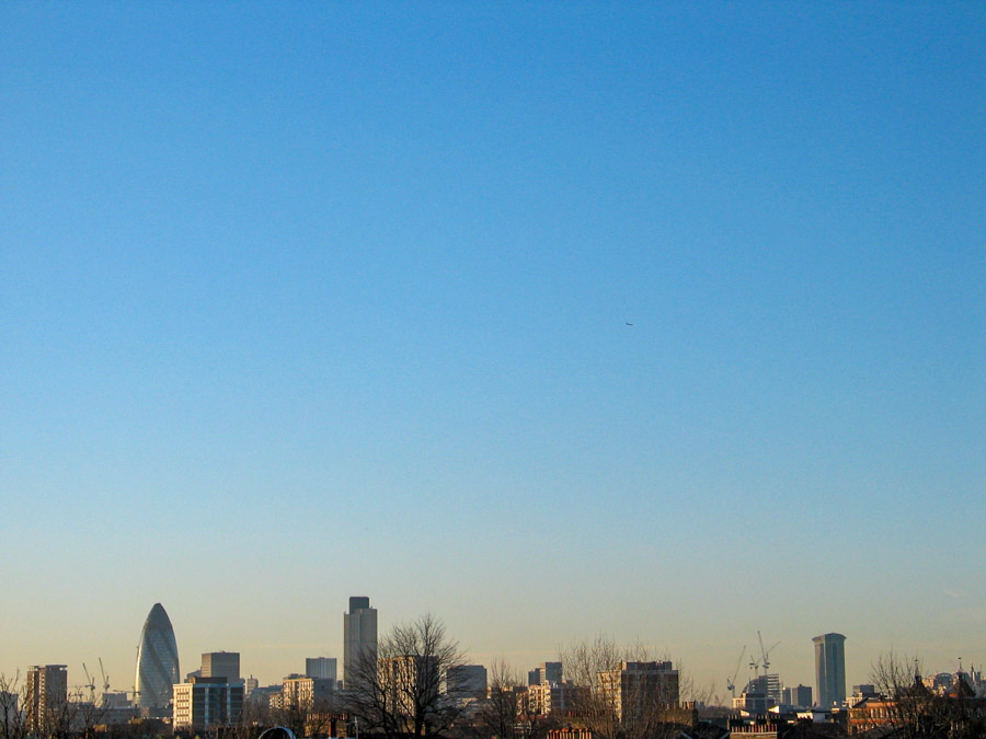 London skyline seen from Hackney under a wide clear blue sky with The Gherkin, Tower 42, and One Canada Square visible above winter rooftops, photographed in 2004 by Antonio Nodar.
