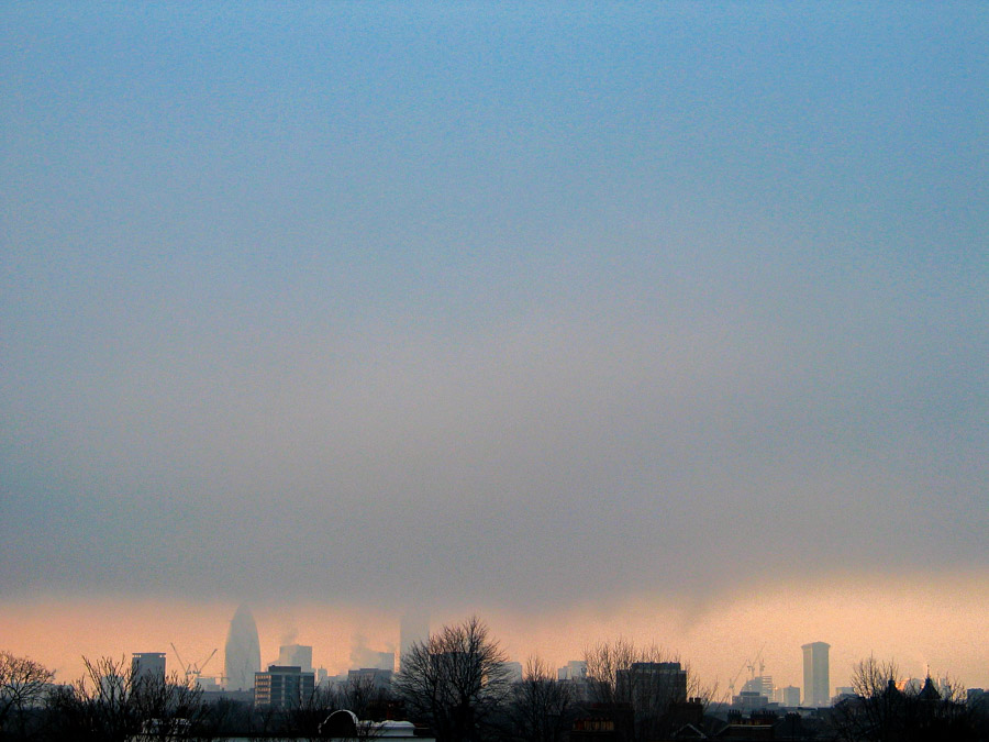 London skyline at dusk seen from Hackney, with The Gherkin, Tower 42, and One Canada Square barely visible behind a thick layer of low cloud and mist above a warm horizon glow, photographed in 2004 by Antonio Nodar.