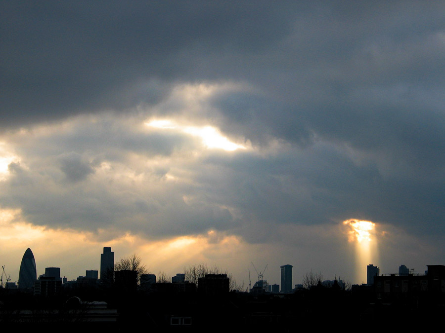Dramatic view of the London skyline from Hackney with The Gherkin, Tower 42, and One Canada Square under dark storm clouds, as bright sunbeams break through openings in the cloud layer, photographed in 2004 by Antonio Nodar.