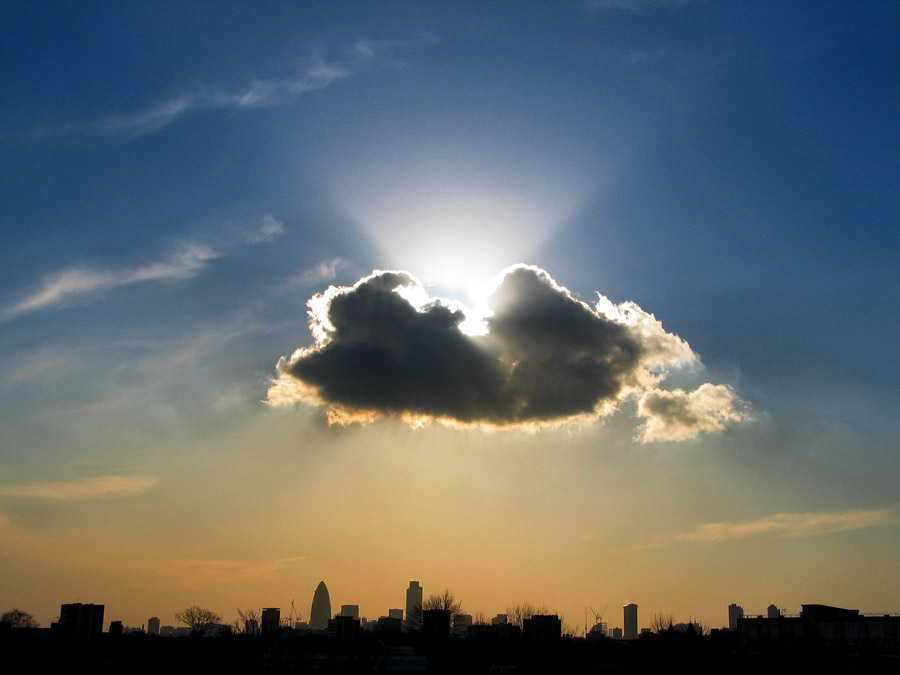 Sunset over the London skyline seen from Hackney, with a bright sunburst behind a single cumulus cloud and The Gherkin, Tower 42, and One Canada Square in dark silhouette, photographed in 2004 by Antonio Nodar.