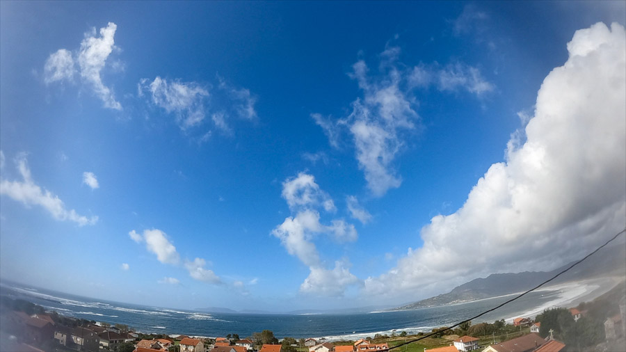 Panoramic seascape of Carnota Bay on the Costa da Morte with fair‑weather cumulus clouds in a bright blue sky above Sofán village rooftops, Monte Pindo, Caldebarcos and Cape Fisterra.