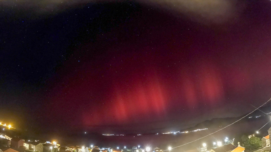 Panoramic night scene over Carnota Bay on Galicia’s Costa da Morte with bright red aurora borealis pillars above the village lights of Sofán, Monte Pindo, Caldebarcos and distant Cape Fisterra.
